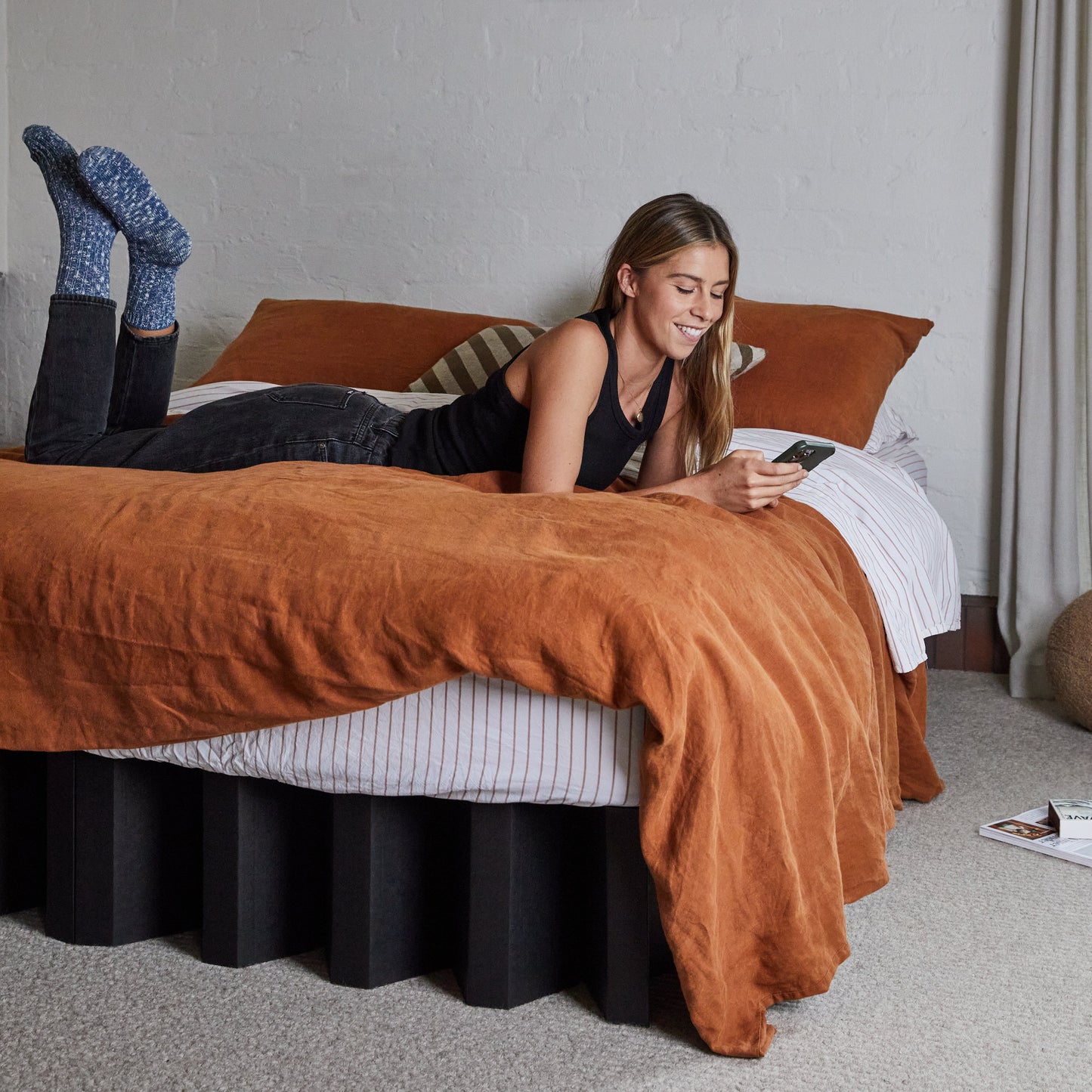 Woman relaxing on Yona Cardboard Bed in black finish with rust-colored bedding and striped pillow in a cozy modern room