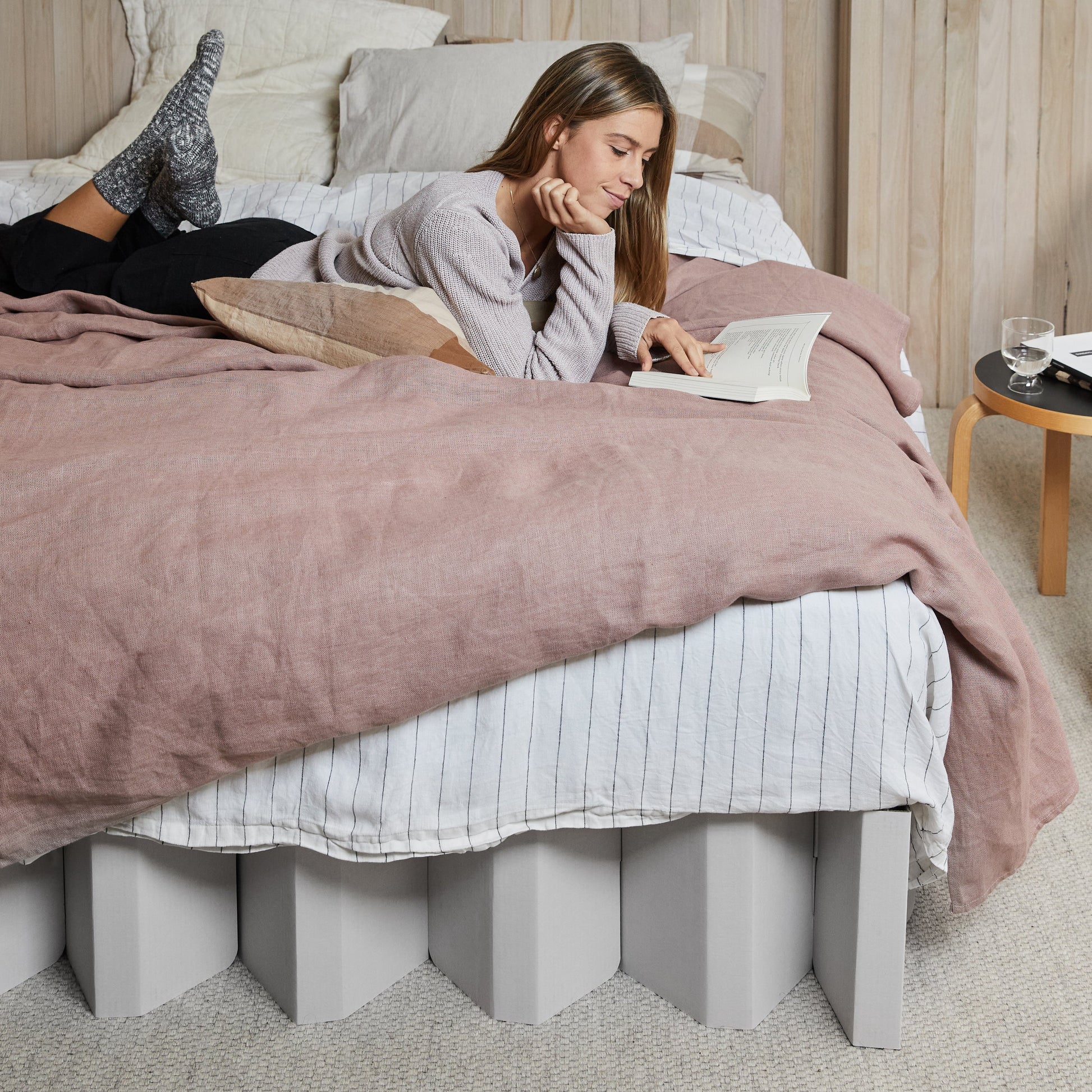 Woman reading on Yona Cardboard Bed in white finish with soft pink bedding and neutral decor